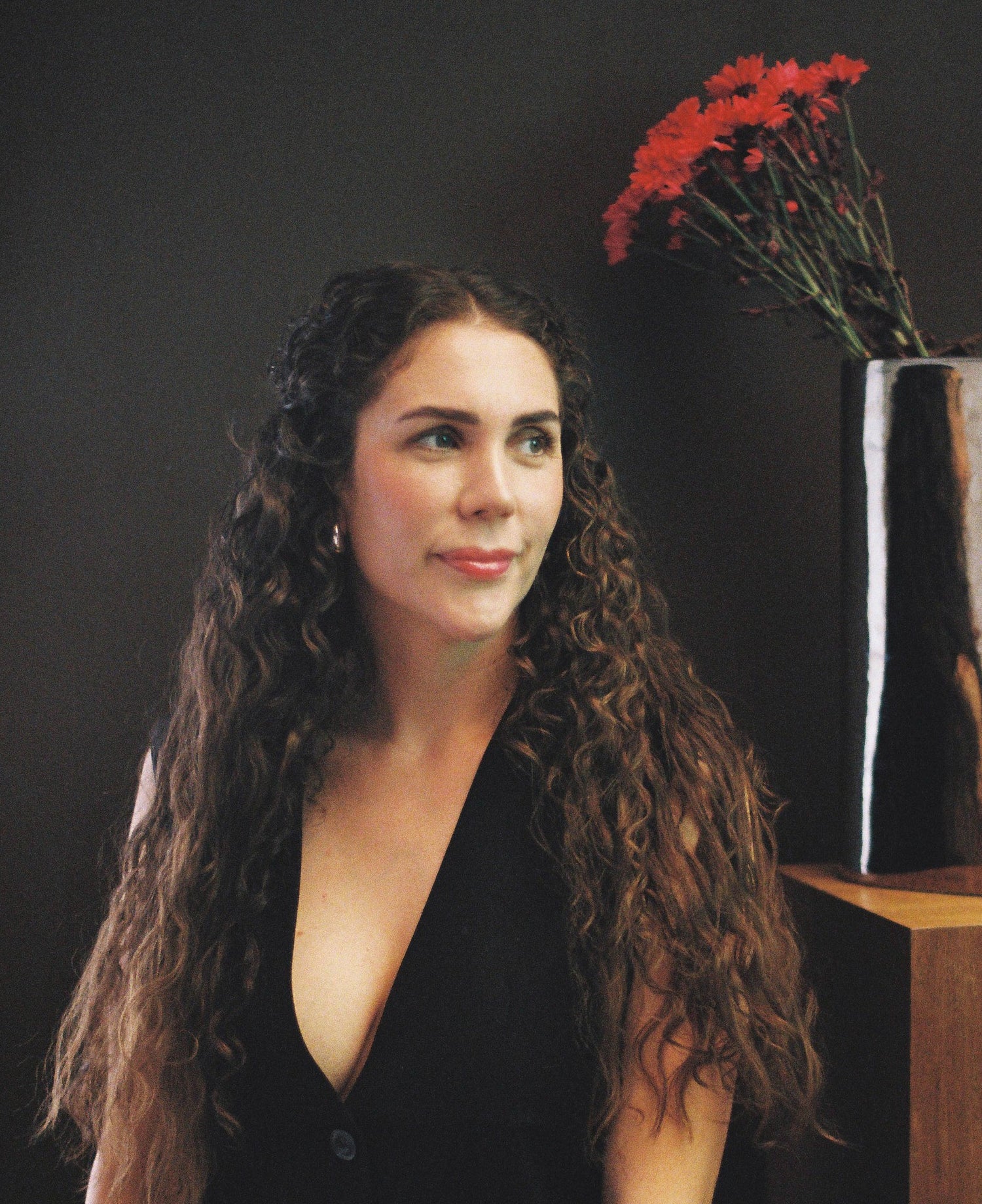 Woman in a black dress sitting next to a silver vase with red flowers on a dark background