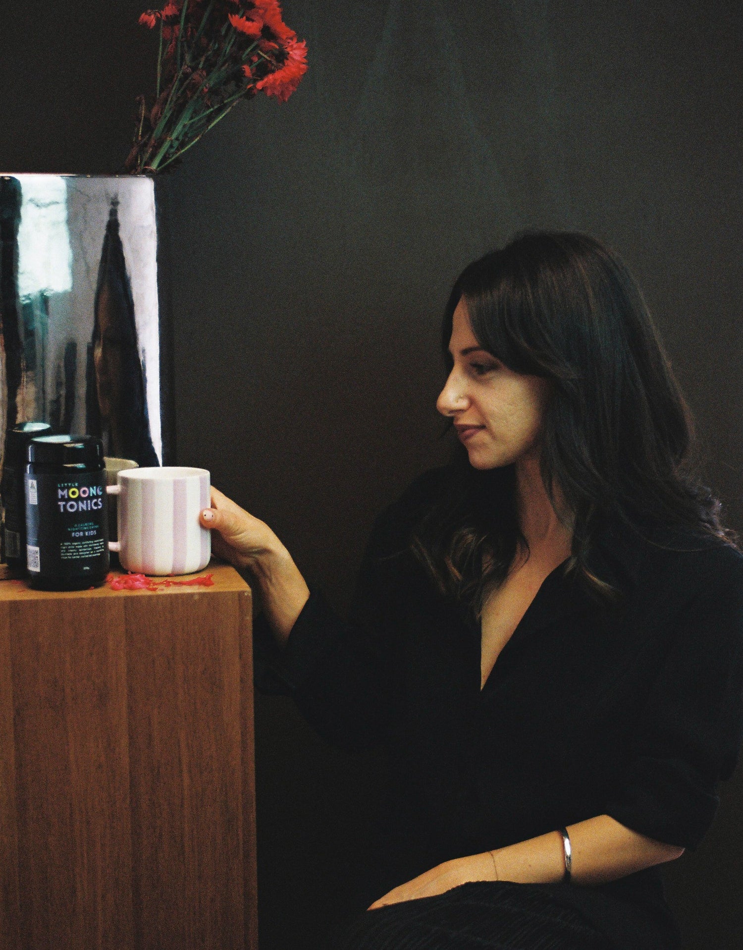 Woman sitting at a table with a cup, surrounded by decor including a vase with red flowers and a canister.