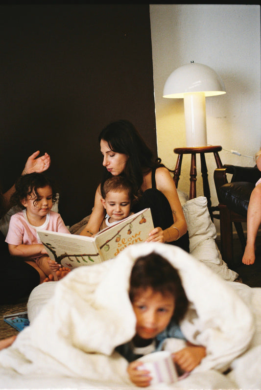 a mother reads a book to 3 children who are sitting beside her while one is snuggled in sheets and drinking a tonic