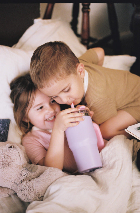 a boy and girl child share a drink from a pastel purple cup with a pink straw