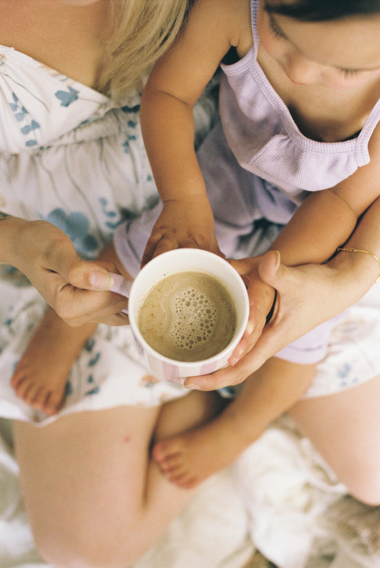 Two children holding a cup of coffee together on a soft surface.