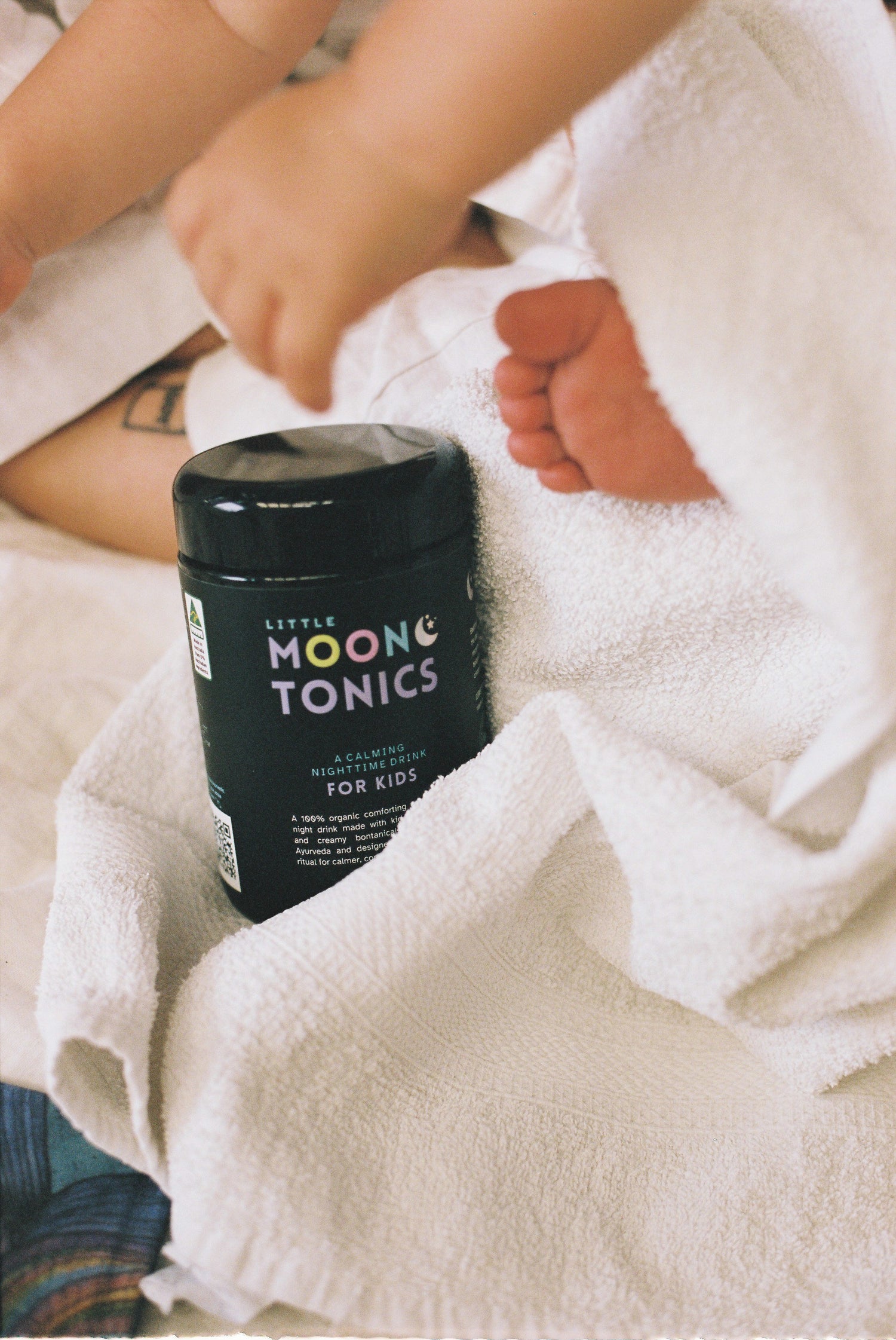 Container of Little Moon Tonics for kids on a white blanket with a child's feet visible.