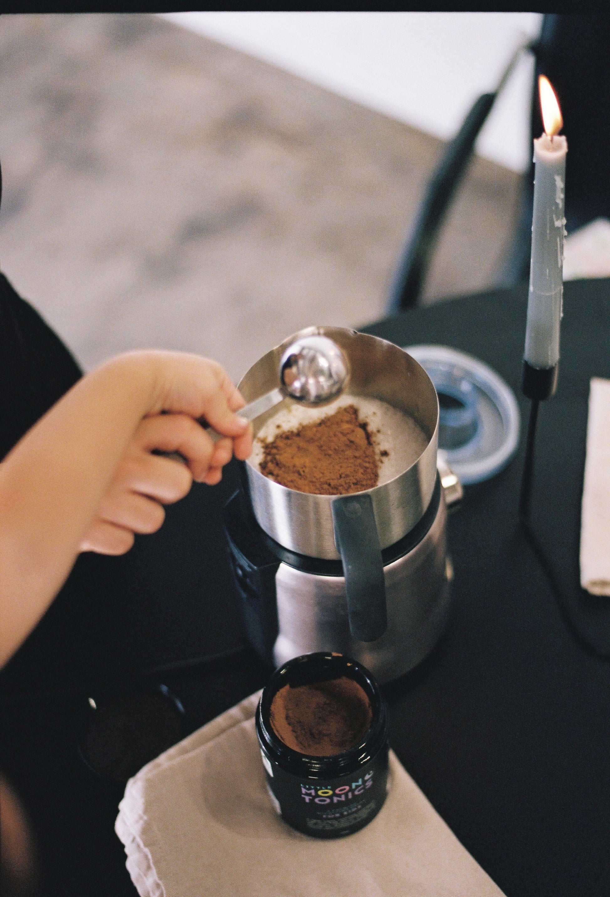 Kid and adult hand pouring scoop of carob-based tonic powder into a stainless steel milk frother full of milk